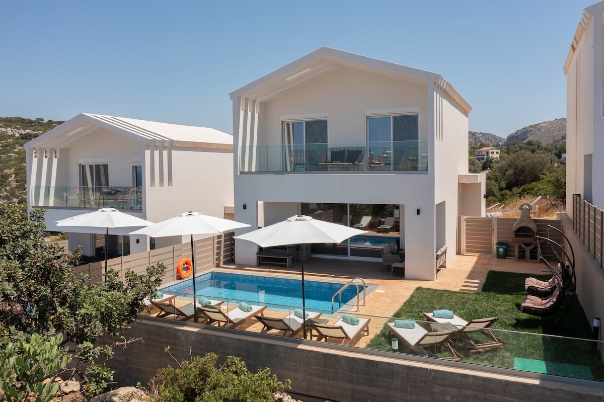 A modern villa exterior is displayed, featuring a private swimming pool surrounded by sun loungers and umbrellas. The upper floor showcases large glass doors leading to private balconies, while landscaped areas enhance the outdoor space. The surrounding hillside completes the tranquil setting.