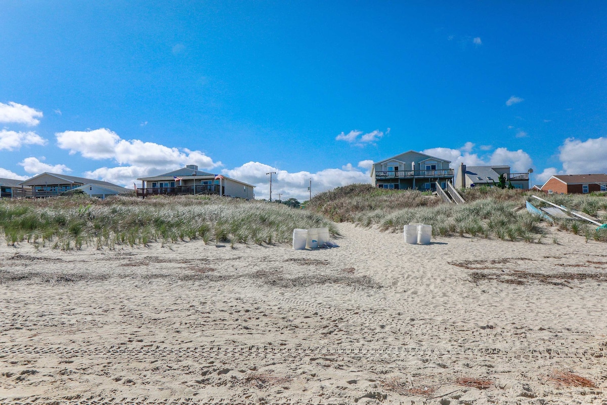 A sandy beach is viewed, leading towards a grassy dune. Residential homes are visible in the background against a clear blue sky. White barrels rest on the sand, indicating beach access and outdoor activities in this coastal neighborhood.