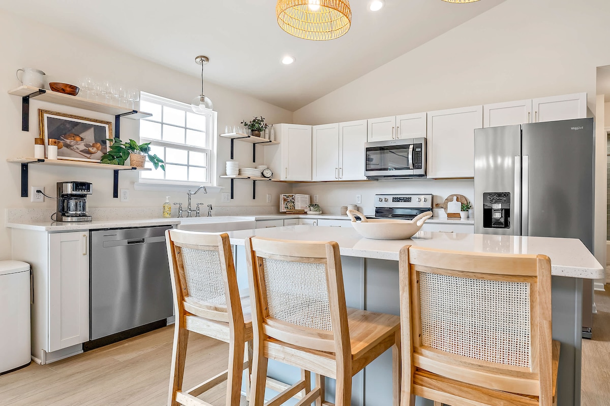 A spacious kitchen features white cabinetry and light countertops, complemented by wooden bar stools with woven backs. Modern appliances, including a refrigerator and microwave, are visible. Natural light streams in through the window, enhancing the clean and inviting atmosphere.