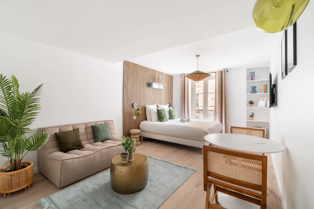 A bright living space features a comfortable sofa, a round dining table, and stylish seating. A spacious bed with a wooden accent wall serves as a focal point. Natural light filters through the curtains, illuminating the decor elements, including plants and bookshelves.
