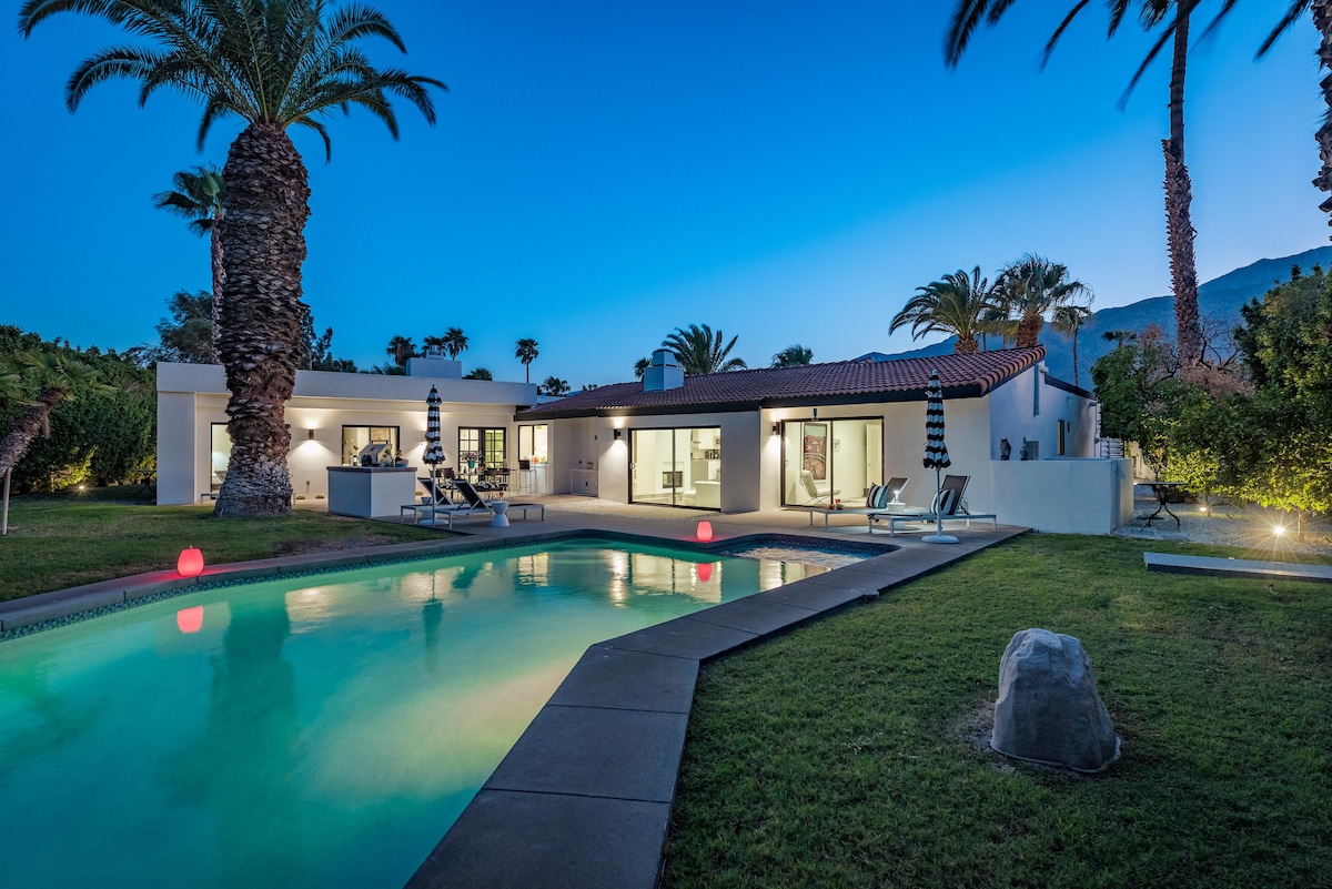 A serene backyard setting features a private swimming pool illuminated softly by floating lights, surrounded by manicured green grass. Palm trees provide ample shade, with a modern home visible in the background. Lounge chairs are positioned near the pool for relaxation.
