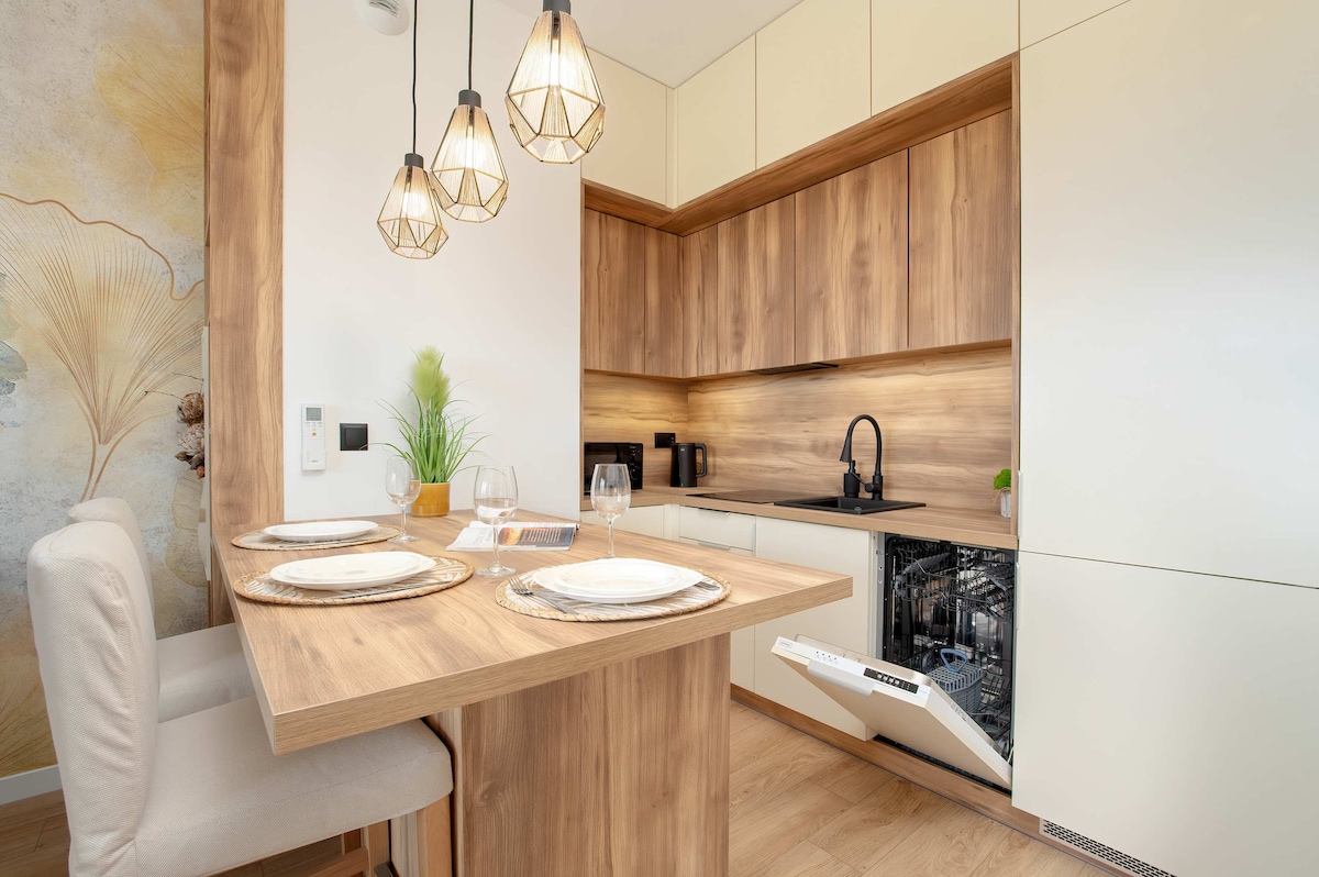 A modern kitchenette features wooden cabinetry and sleek appliances, including a dishwasher. The small dining area is set with a table for two, complemented by decorative plates and wine glasses. Three pendant lights add a warm glow to the inviting space.