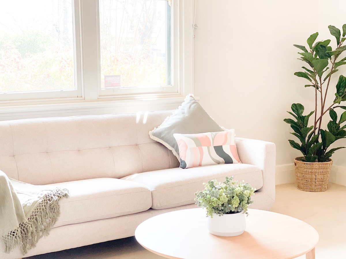 A light-filled living area features a plush, cream-colored sofa adorned with decorative cushions and a soft throw. A round coffee table sits before the sofa, topped with a small potted plant. A green indoor plant is positioned in a woven basket, adding a touch of nature.