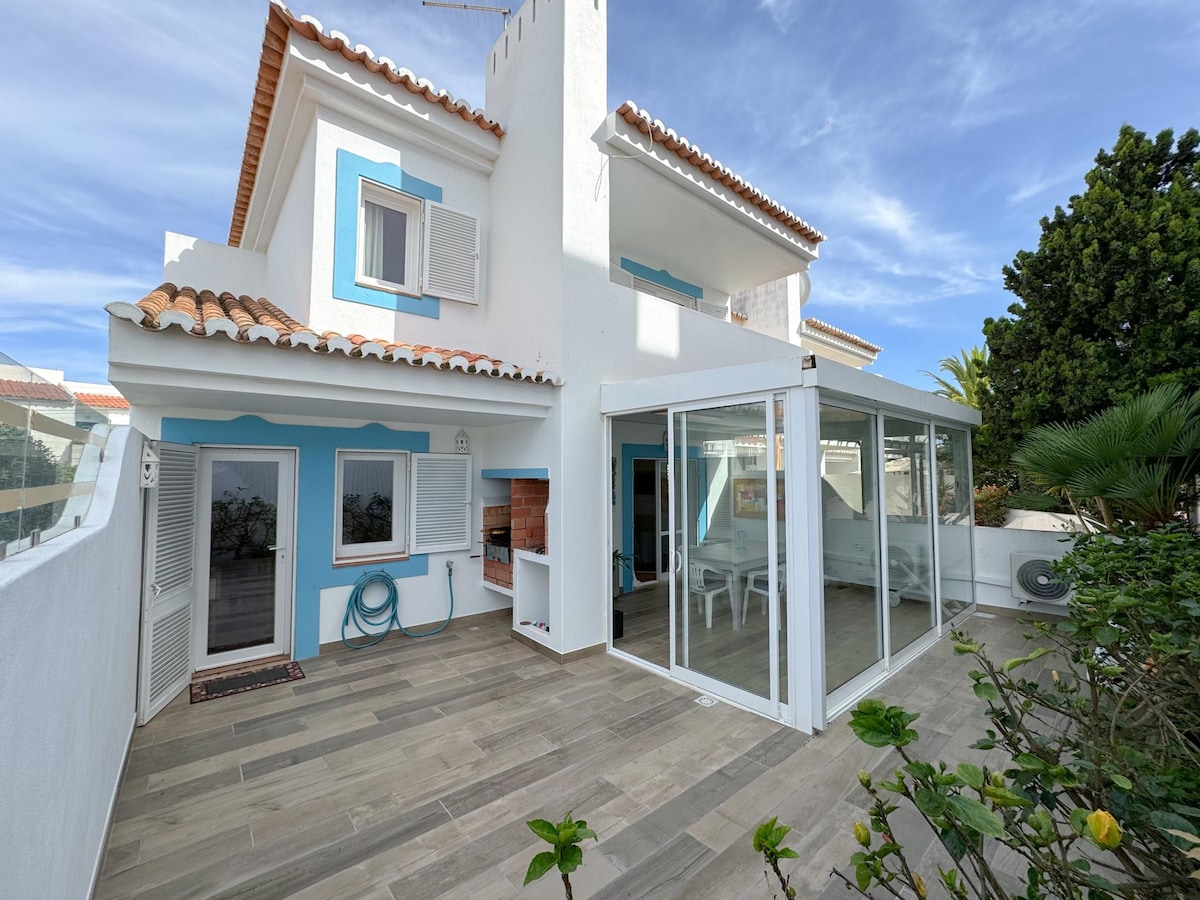 The exterior of the villa features white walls with blue accents, showcasing a sunny patio area. A glass-enclosed space provides a shaded setting for relaxation. The outdoor area is complemented by surrounding greenery, enhancing the inviting atmosphere of the home.