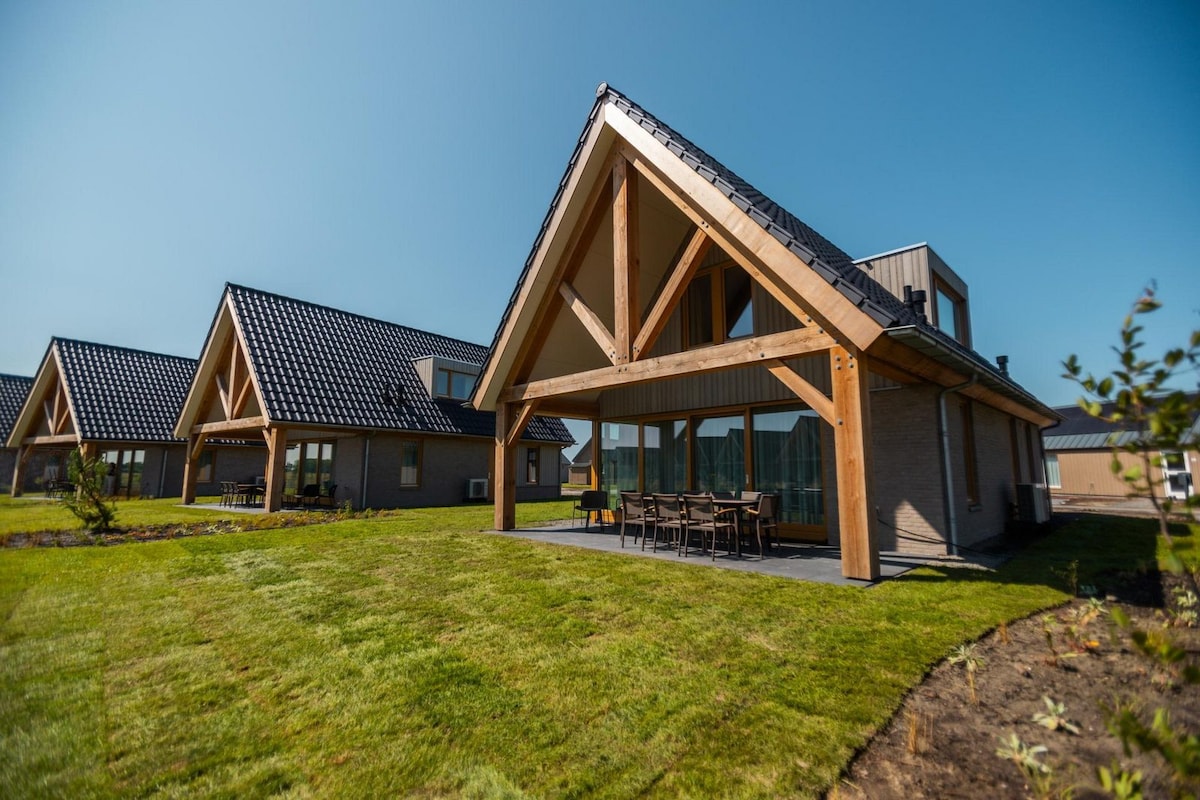 A modern villa is showcased with a sloping roof and large windows, surrounded by well-maintained grass. The outdoor dining area features a long table with multiple chairs. Nearby structures exhibit similar architectural styles, enhancing the cohesive look of the property.