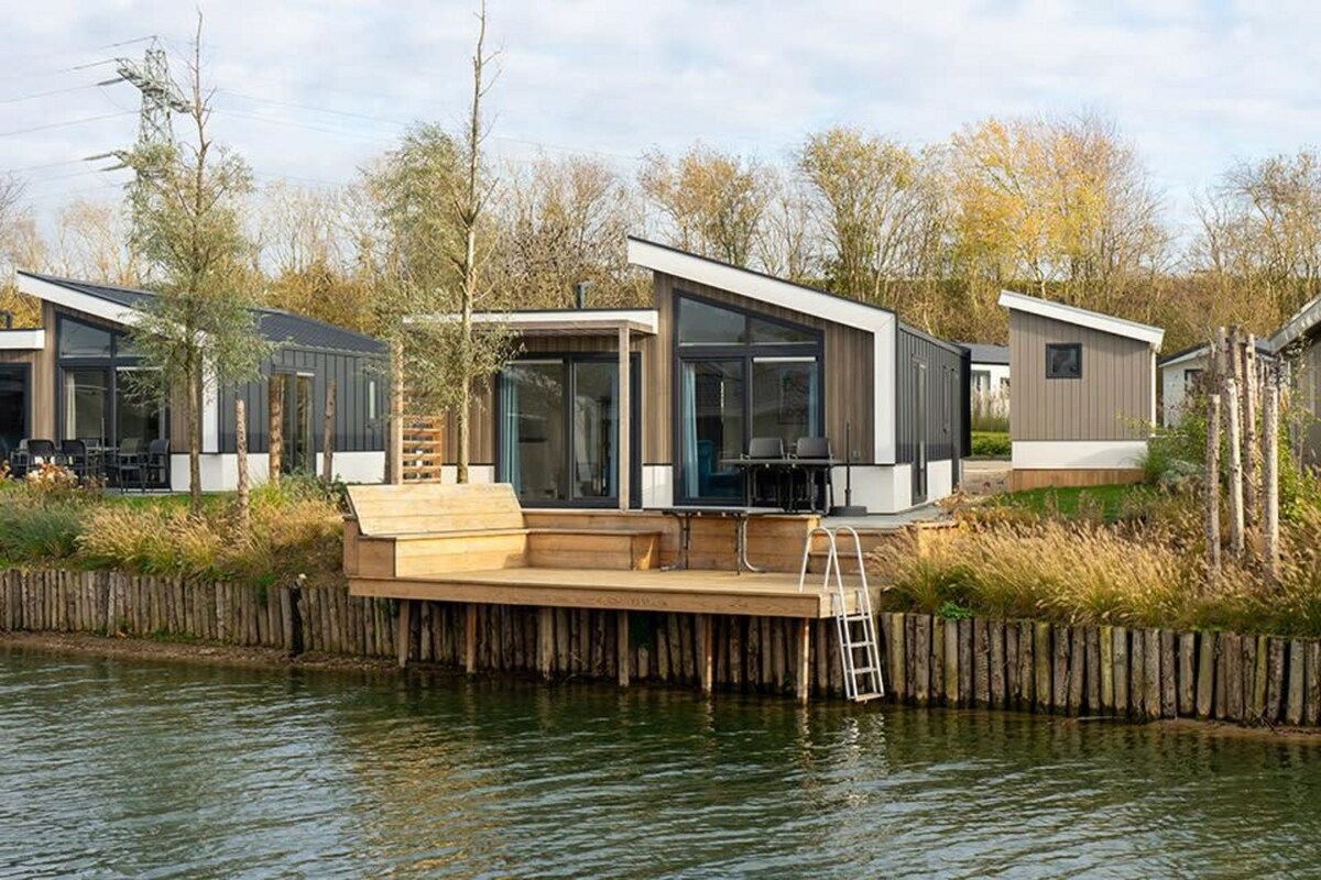 A modern chalet is depicted alongside a serene waterfront, featuring large glass windows and a slanted roof. A wooden jetty extends from the building, complemented by outdoor seating. Lush grass and trees border the property, contributing to the tranquil setting.