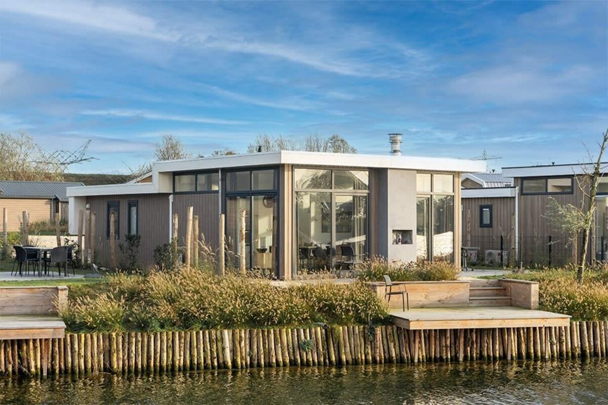 The exterior of a contemporary detached waterfront chalet is showcased, featuring large windows that allow natural light to fill the interior. The structure has a round design, surrounded by grassy areas and wooden decking along the water's edge, with a clear blue sky above.