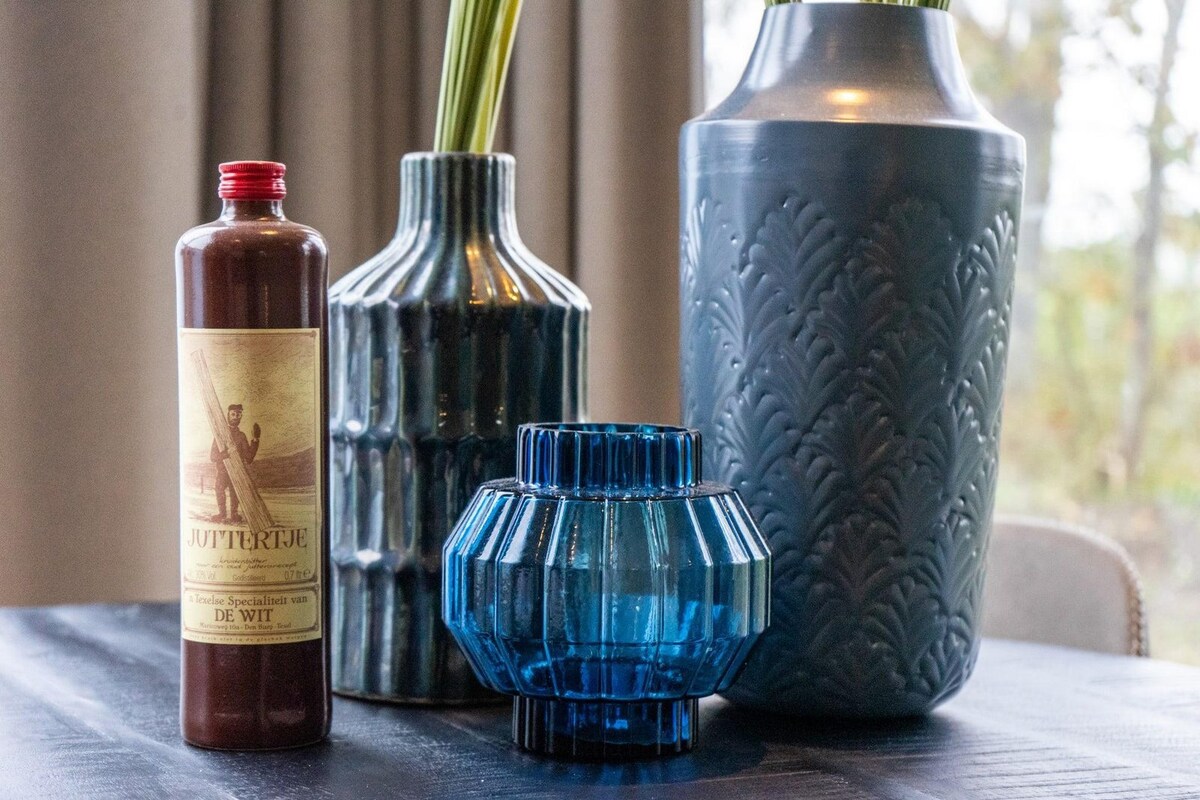A collection of decorative vases and a bottle is displayed on a dark surface. The vases vary in shape and texture, featuring a blue glass vase, a ribbed ceramic vase, and a green-toned ceramic vase. A vintage-style bottle adds contrast to the arrangement.
