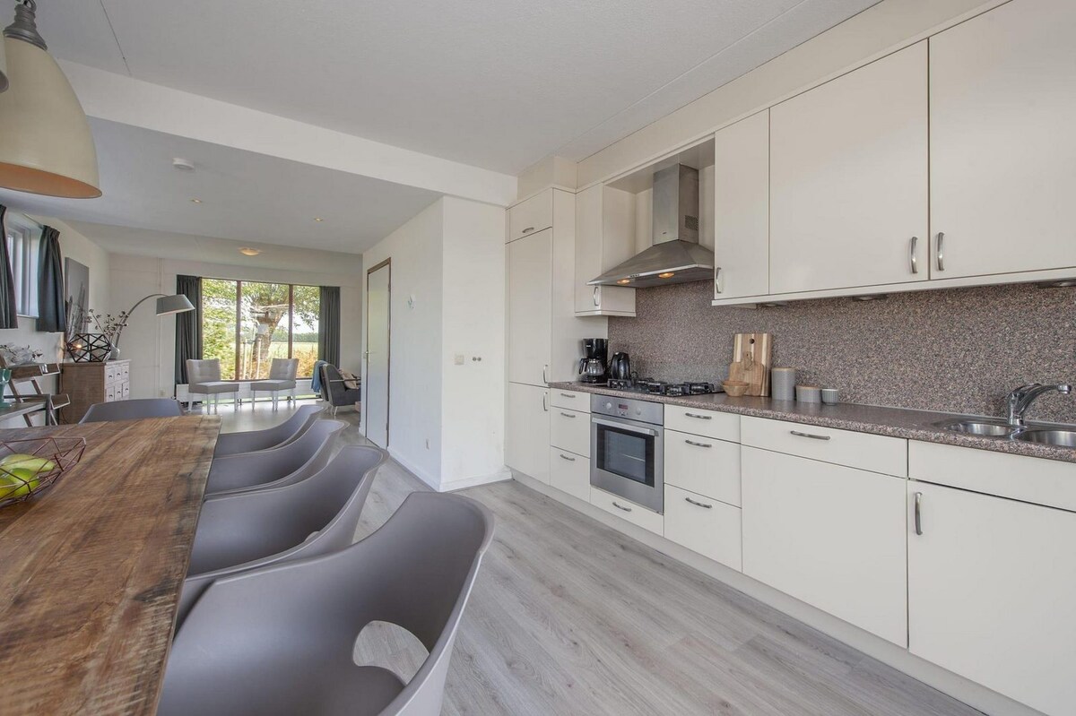 An open kitchen is featured, showcasing light-colored cabinetry and modern appliances including a stove, oven, and refrigerator. A wooden dining table is accompanied by four sleek chairs. Natural light fills the space from large windows leading to an outdoor area.