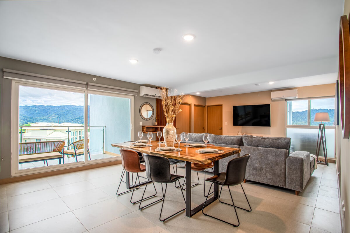 A modern dining area features a wooden table set for six beside an inviting sectional couch. Large windows provide natural light and mountain views, while wall decor adds subtle elegance. A television is mounted on the wall, complementing the contemporary design.