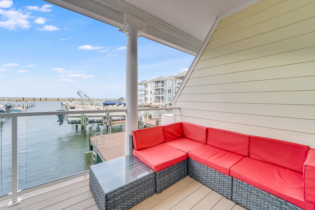 A waterfront balcony features a spacious outdoor seating area with red cushions and a glass-topped table. The view overlooks the marina, showcasing boats docked along the pier against a backdrop of clear blue skies.