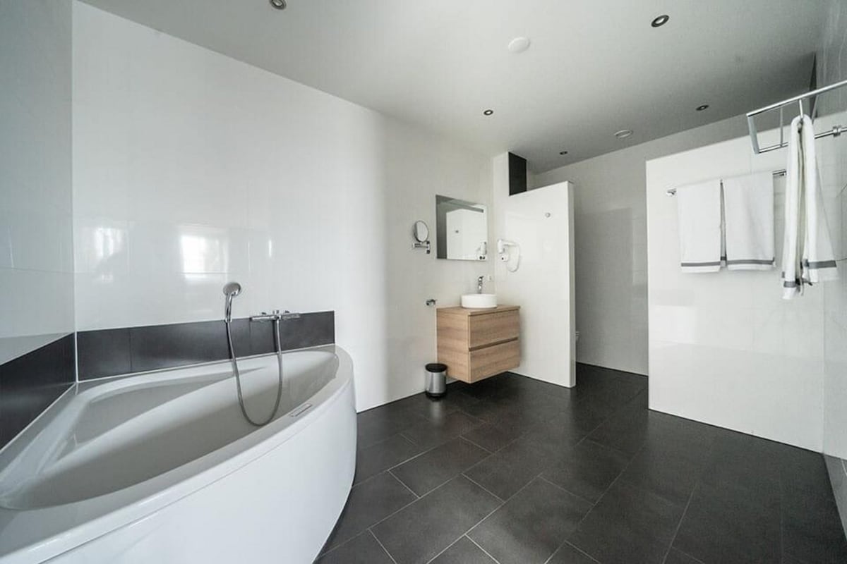 The spacious bathroom features a modern, curved bathtub alongside a sleek wooden vanity. A large mirror is mounted above the sink, while white towels are neatly hung on a rack. The minimalist design is complemented by dark, tiled flooring and bright, neutral walls.