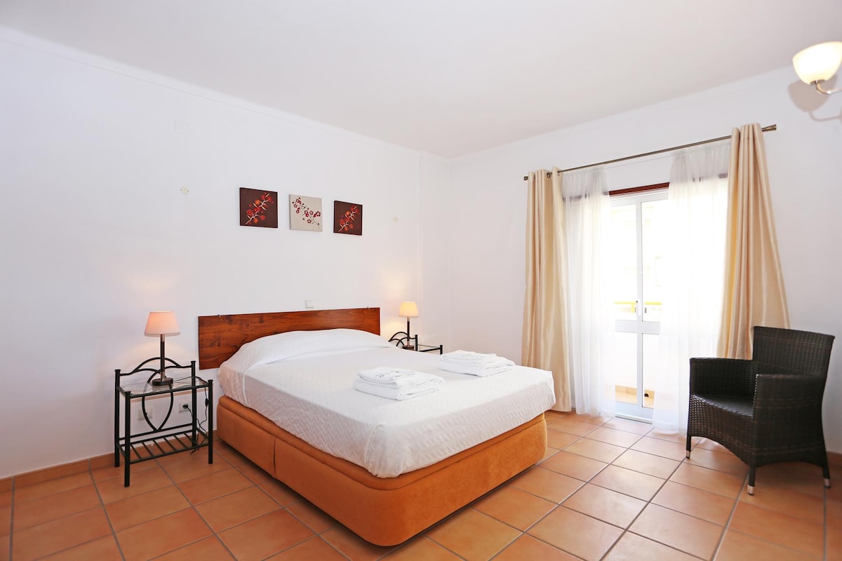 The private bedroom is designed with a double-sized bed and equipped with fresh linens. Two bedside lamps illuminate the space. A chair is positioned beside the window, which is dressed with light curtains, allowing natural light to enter. Wall art adds a touch of decoration.