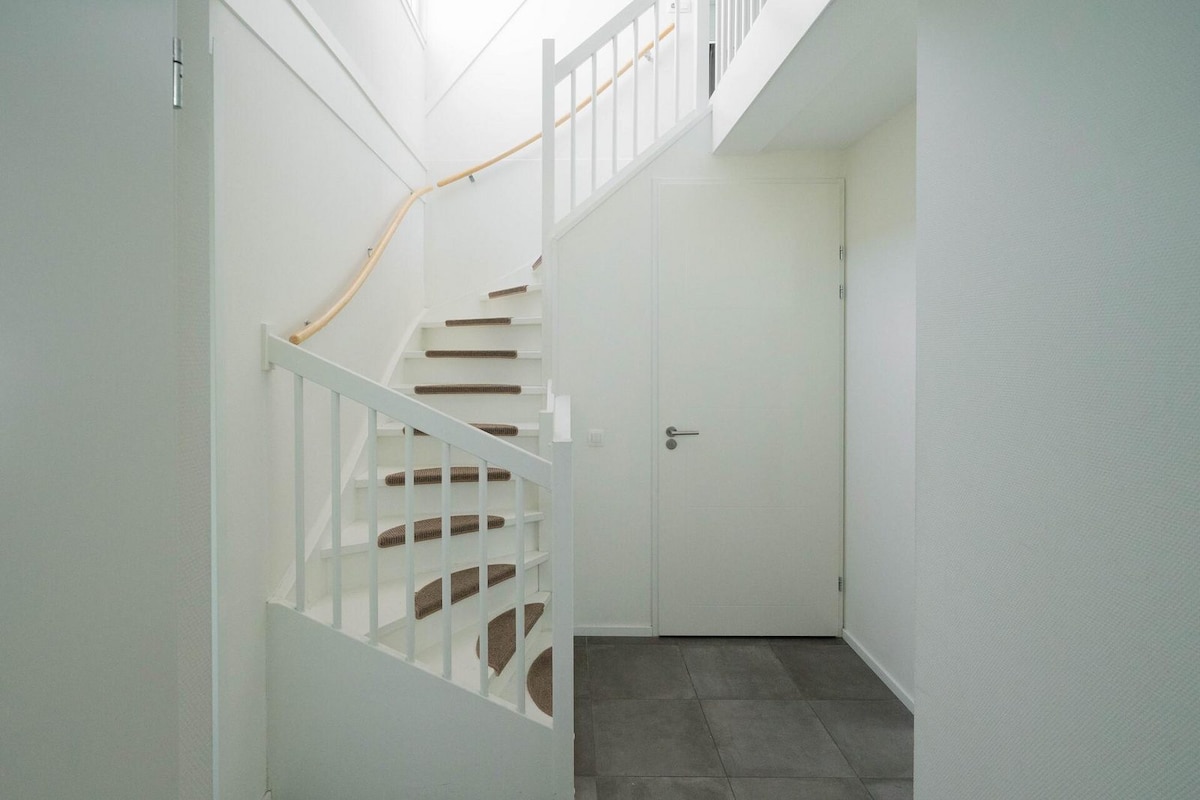 A well-lit entrance features a staircase with a wooden handrail, leading to the upper floor. The walls are painted in light tones, complemented by a spacious tiled area at the base of the stairs. A closed door is visible beside the stairway.