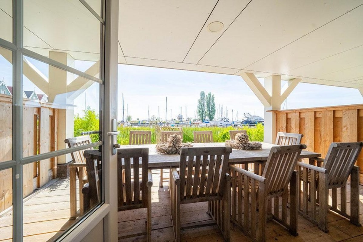 A spacious terrace is furnished with a large wooden table surrounded by multiple chairs. The area offers a view of the marina, showcasing boats and lush greenery in the backdrop. Natural light brightens the space through an open door.