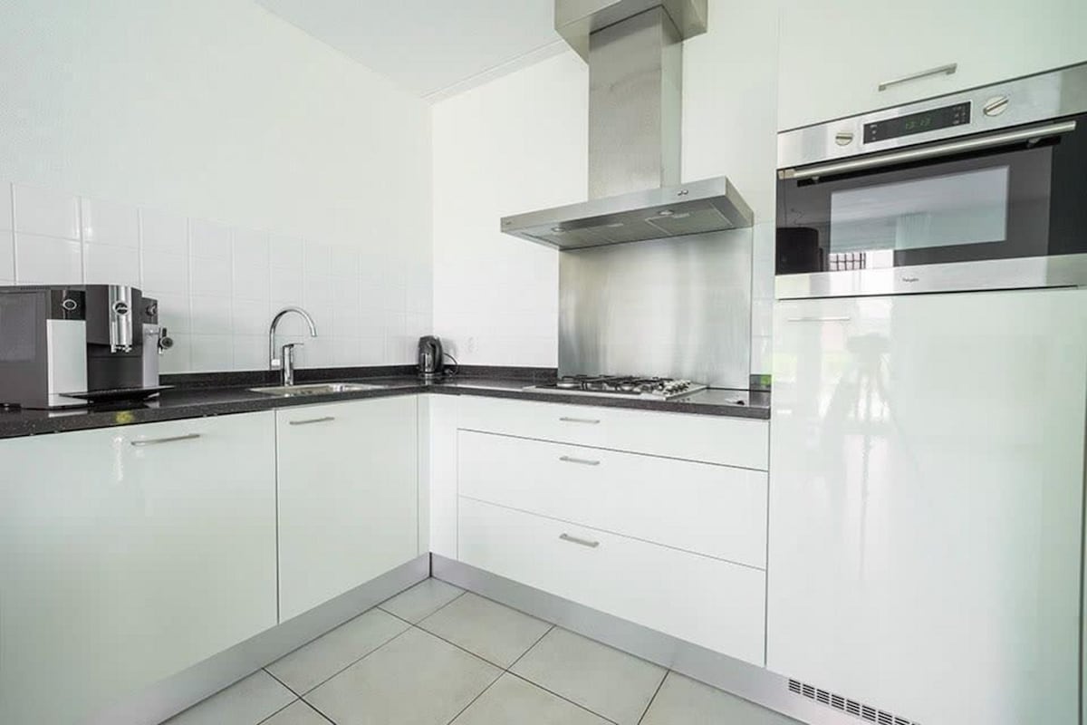 A modern kitchen is presented with sleek white cabinetry and a black countertop. Appliances include a built-in microwave, an oven, a stainless steel range hood, a gas cooktop, and a coffee machine. The area is well-lit, with tiled flooring enhancing the clean design.