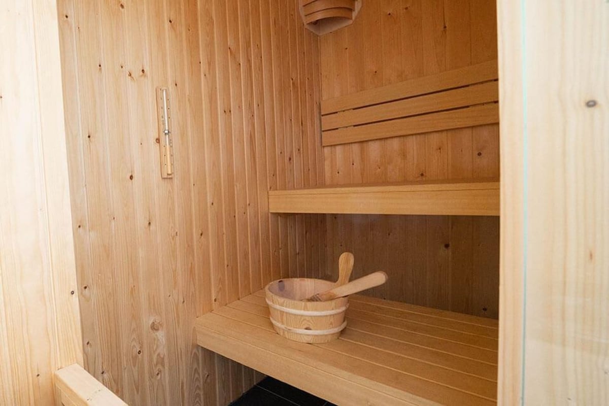A traditional sauna is featured with wooden paneling, creating a warm environment. Bench seating is provided on two levels, with a wooden bowl resting on the lower shelf. Subtle lighting enhances the natural wood tones, offering a serene space for relaxation.