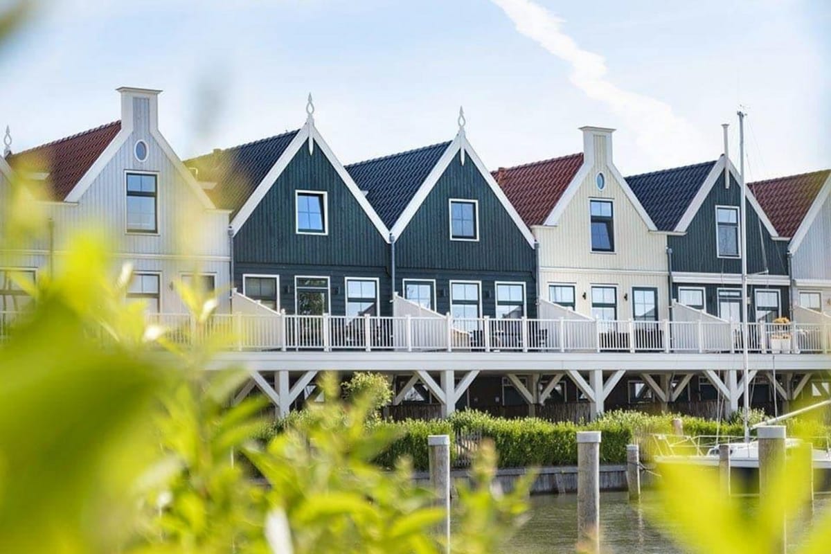 A row of charming flat exteriors is visible, featuring a mix of colors, including green and beige. Each flat has a balcony with white railings and outdoor seating. The setting is enhanced by greenery in the foreground and water in the background, suggesting a coastal environment.