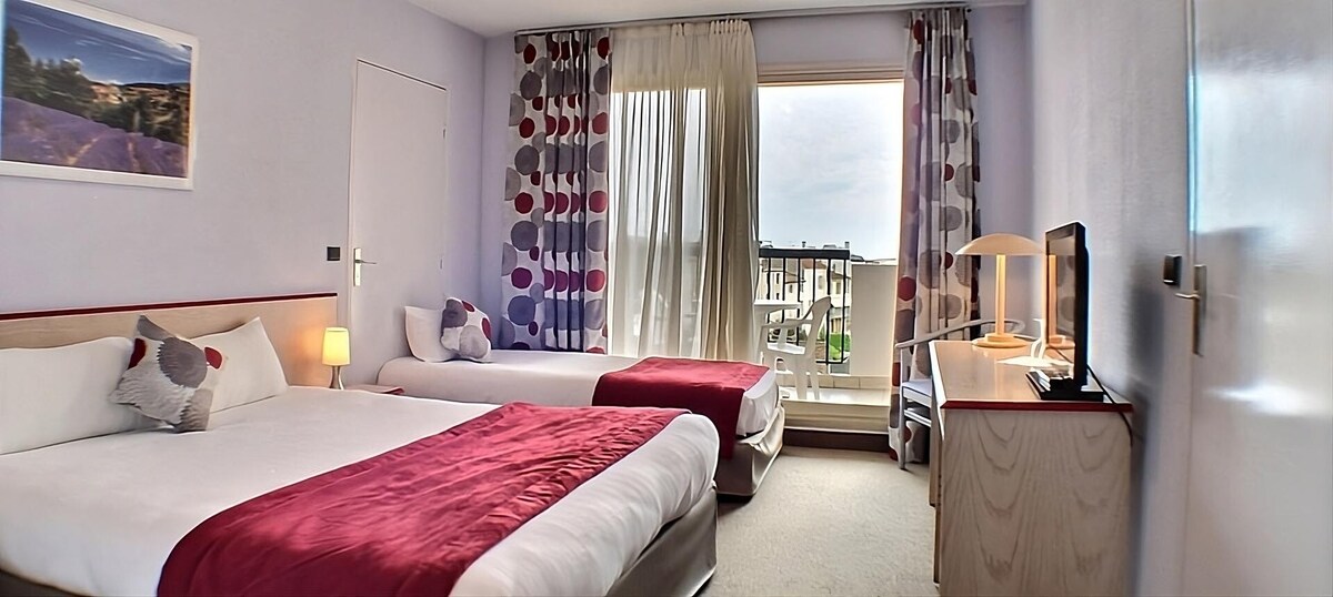 A comfortable room features a double bed and a single bed, accompanied by a flat-screen TV and a desk with a chair. Large windows allow natural light to fill the space, which is adorned with contemporary curtains and artwork.