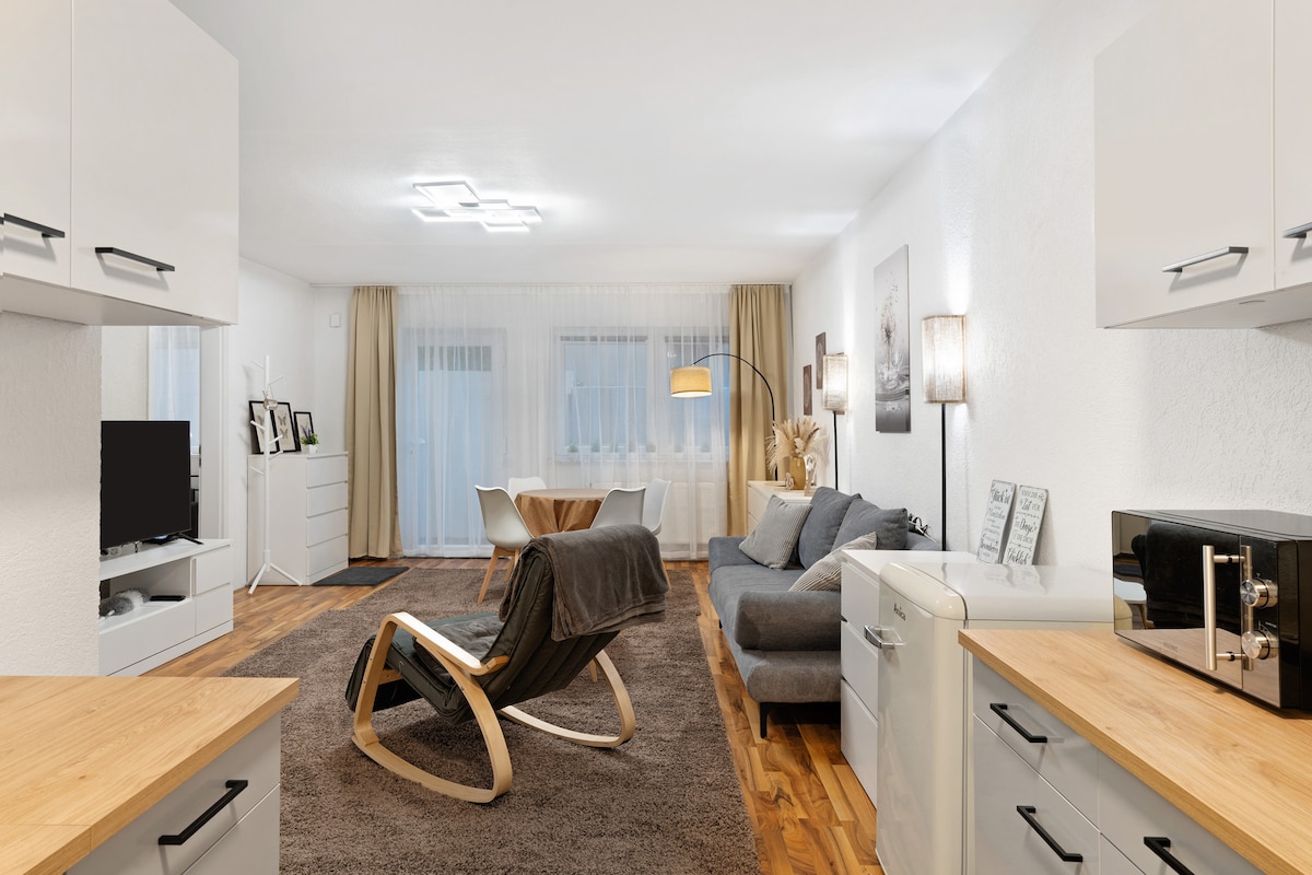 A modern living area is presented with a sleek grey sofa and a wooden rocking chair. Soft lighting is provided by floor lamps. A dining table set for two is visible in the background, next to large windows draped with sheer curtains. Warm wooden flooring complements the space.