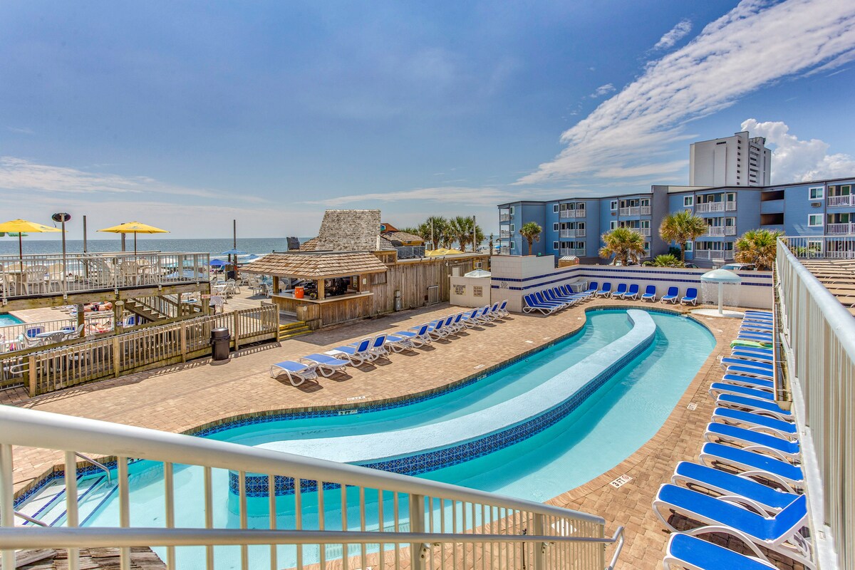 The outdoor pool area features a winding pool surrounded by lounge chairs. The pool's serene blue water reflects the sky, with beach access visible in the background. Palm trees provide a tropical feel, complementing the nearby resort buildings.
