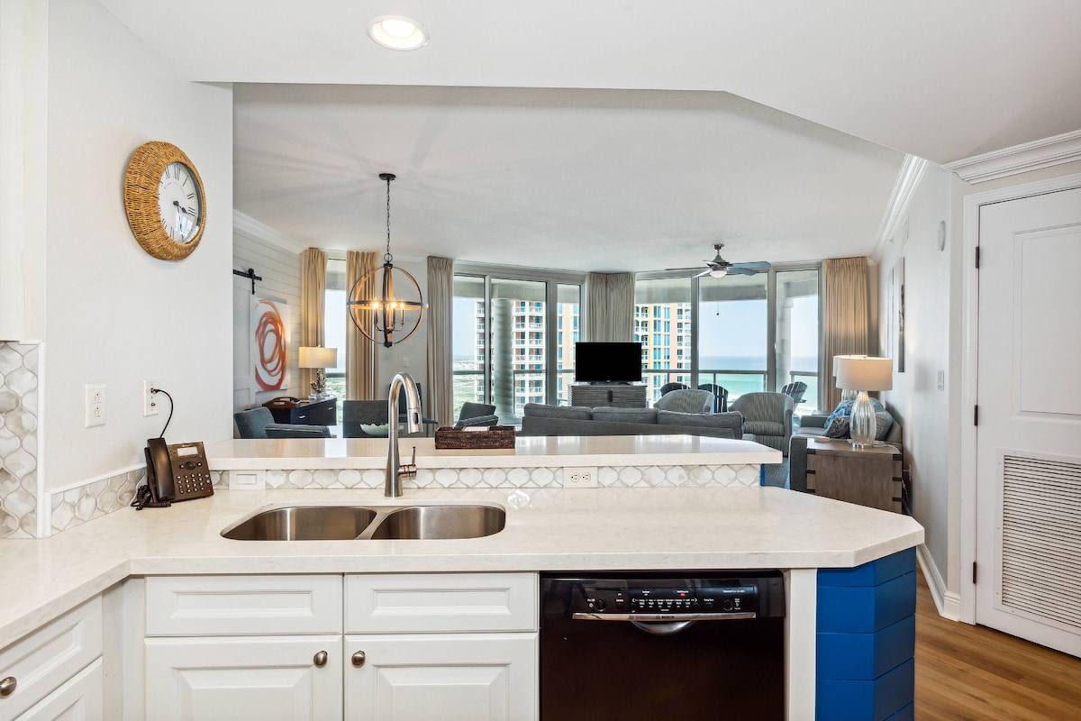 The kitchen features a modern design with white cabinetry and a light countertop. A double sink is prominent, while the view extends into the living space, highlighted by floor-to-ceiling windows that provide natural light and views of the seating area.