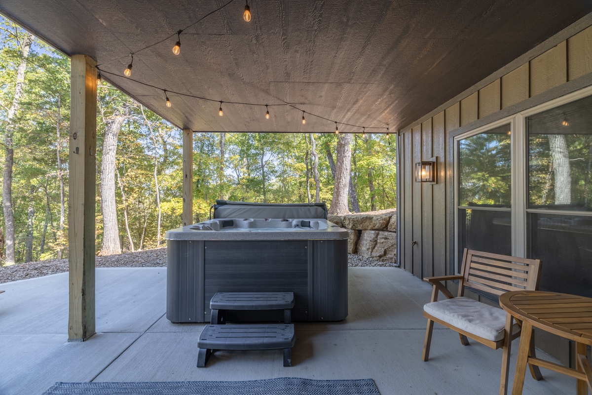 A covered outdoor space features a spacious hot tub, surrounded by warm lighting from string lights overhead. A small table and a chair provide a cozy seating option, with views of the wooded area visible through large windows.
