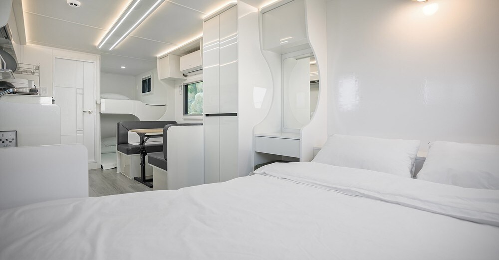 A modern studio room features a spacious bed with white bedding, accompanied by a seating area with a table and chairs. Bright, clean lines and neutral tones create an open atmosphere, with natural light flowing in through the window.