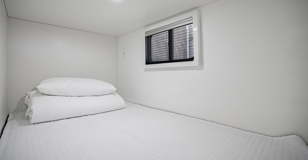A compact sleeping area is featured, with a snug mattress covered in white bedding. Two fluffy pillows are neatly arranged at the head of the bed. A small window allows for natural light, while white walls create a clean, minimalist aesthetic.