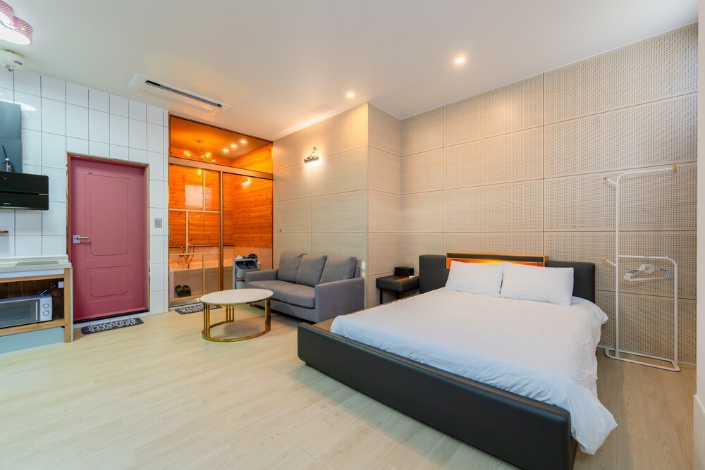 A spacious room with light-toned wood flooring features a large black bed with crisp white linens. A grey sofa is positioned nearby, complemented by a circular coffee table. The kitchenette area includes modern appliances, and an adjoining room is visible in the background.
