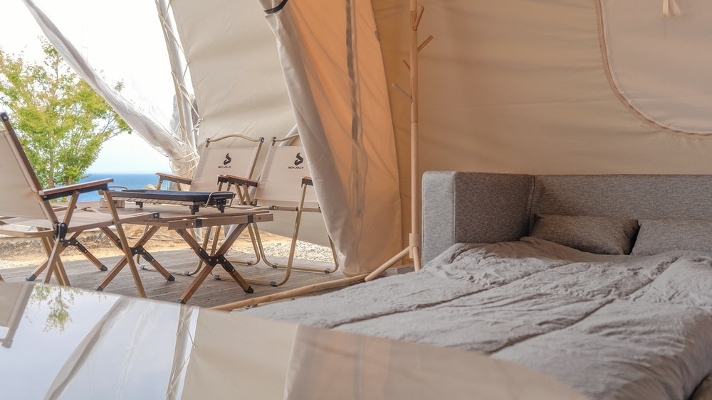 A spacious tent interior is displayed, featuring a cozy bed with soft bedding. Two folding chairs and a table are positioned just outside, with a view of greenery and the sea visible through the entrance.