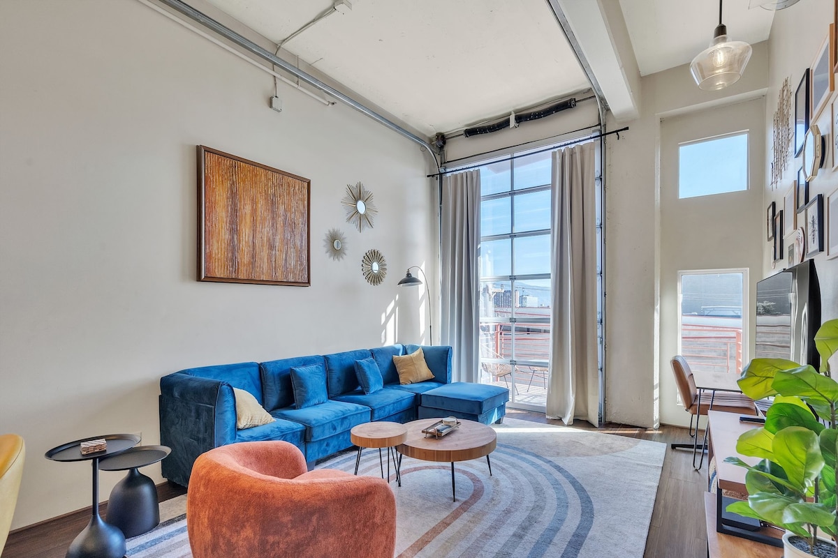 A spacious living area features a large blue sectional sofa complemented by contemporary accent chairs. Natural light fills the room through large windows, framed by light curtains. A wooden coffee table rests on a patterned rug, with wall art providing visual interest.