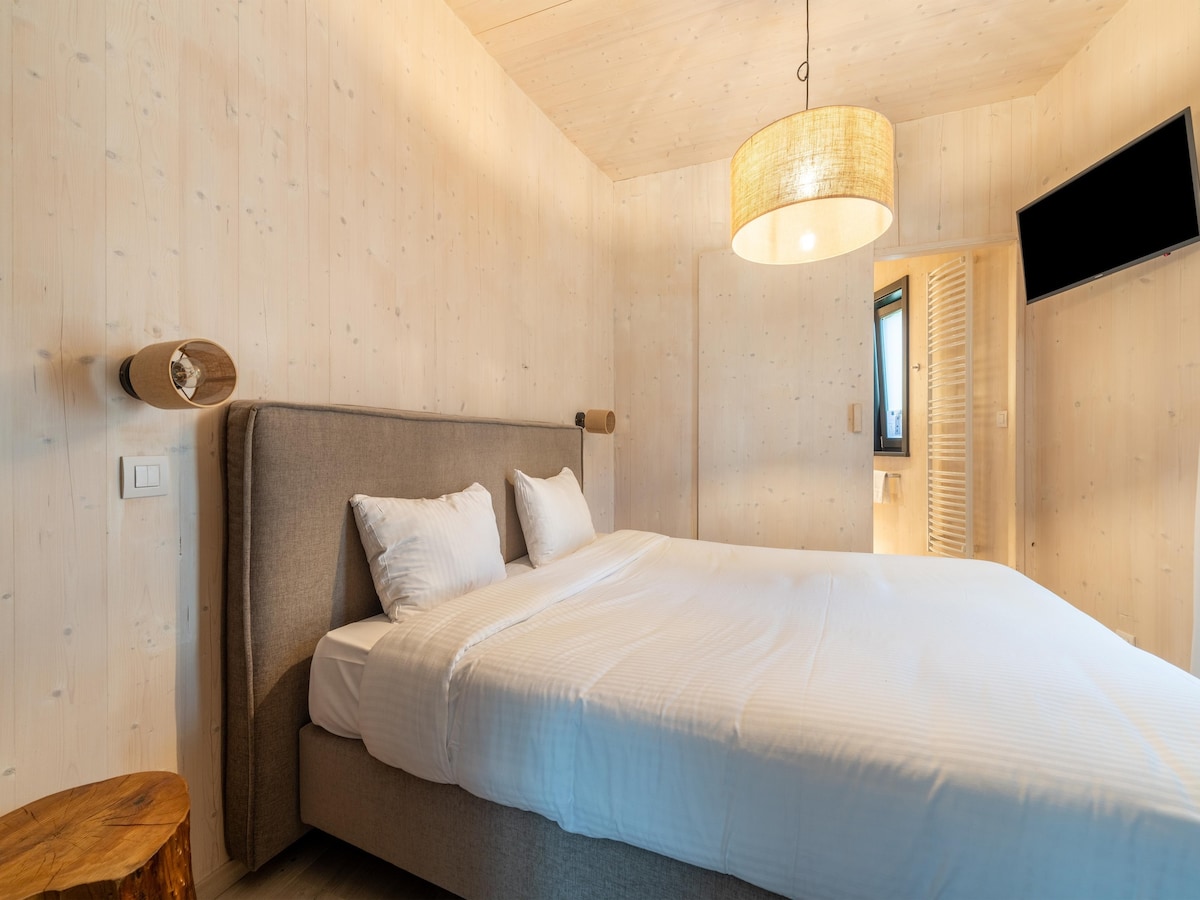 A cozy bedroom features warm wooden walls and a plush double bed with crisp white linens. Soft lighting is provided by a pendant lamp overhead, while a wall-mounted flat-screen TV is positioned for viewing convenience. A rustic side table is present beside the bed.