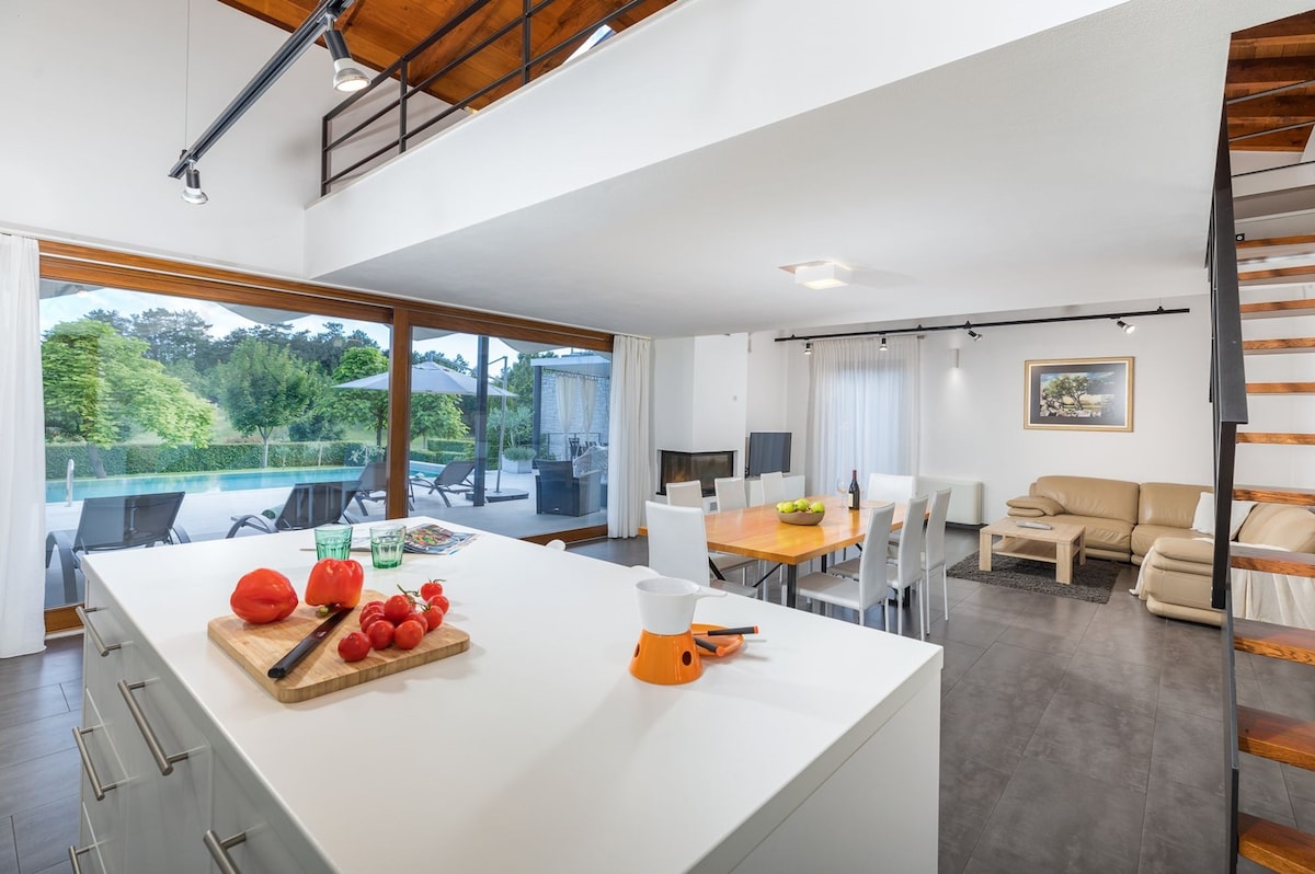 An open-plan kitchen and dining area features a central island with fresh vegetables and utensils. A dining table accommodates multiple guests, while a cozy living area includes a sofa nearby. Large glass doors offer views of the outdoor terrace and pool area.