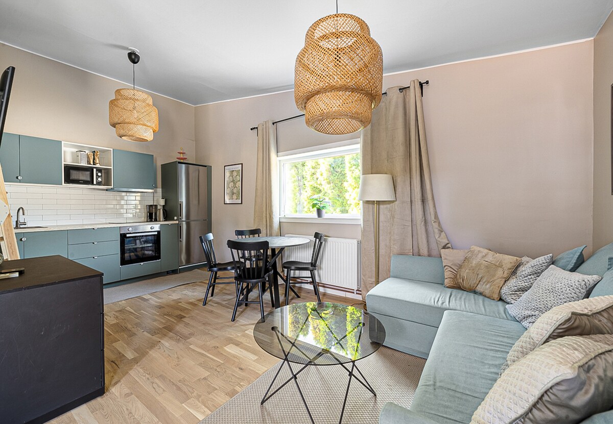A modern kitchen and living area is depicted, featuring a combination of light blue cabinetry and white tiled backsplash. A dining table for four is positioned next to the large window, which is dressed with natural curtains. A comfortable sectional sofa and woven light fixtures enhance the laid-back ambiance.
