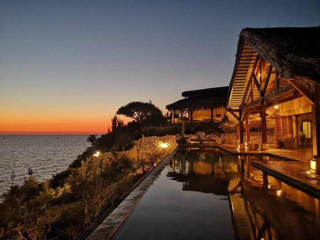 An inviting infinity pool stretches along the edge of a coastal property, reflecting the gradient hues of a sunset over the ocean. Natural wood elements of the structure complement the surrounding gardens. Soft lighting provides a warm glow as night approaches.