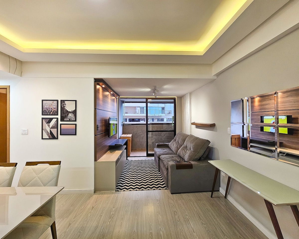 A modern living area features a gray sectional sofa facing a large window, allowing natural light to fill the space. A sleek dining table is seen to the left, with minimalist decor showcased on the walls. Soft lighting highlights the room's clean lines.