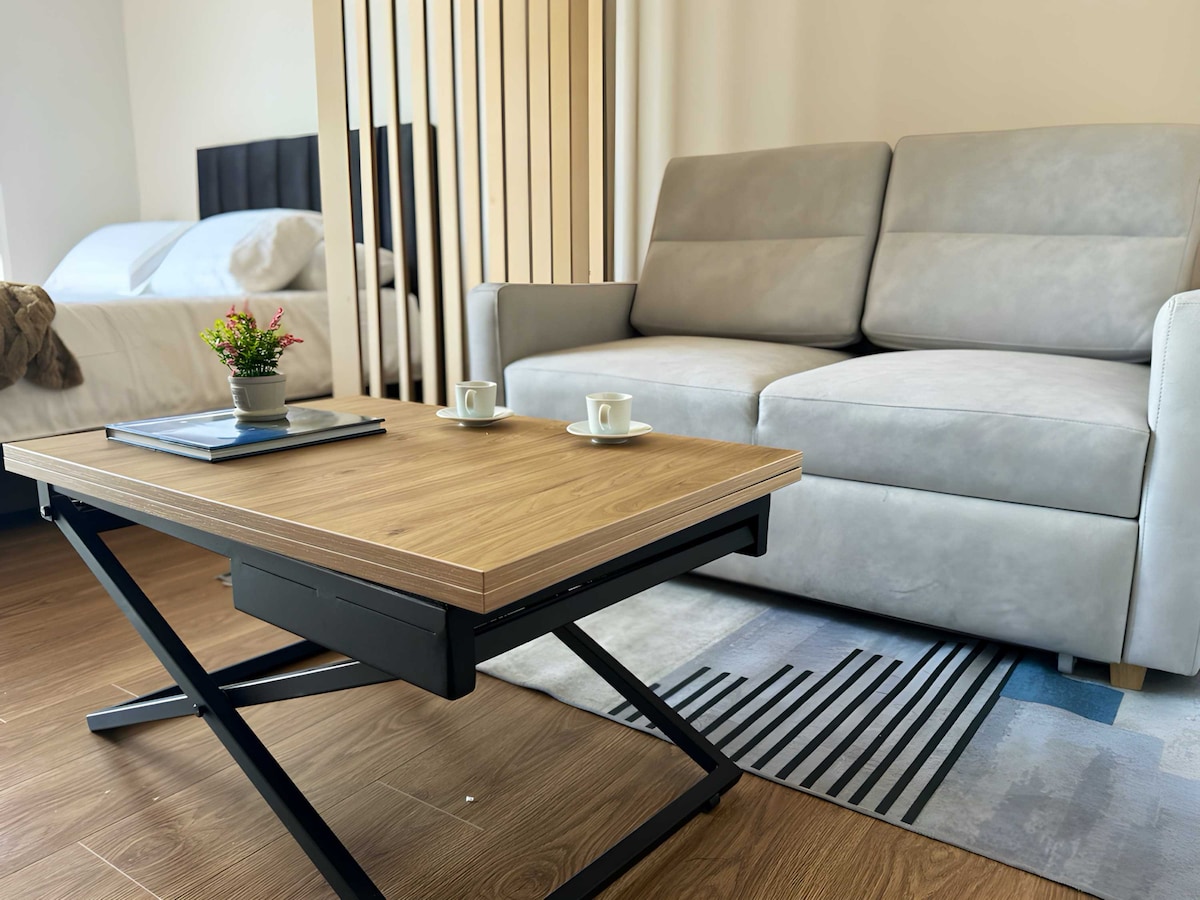 A multi-purpose table is positioned in front of a comfortable sofa, featuring a wooden surface and a minimalist design. Nearby, a small plant adds a touch of greenery. The backdrop includes a neatly made bed, creating a functional living space.