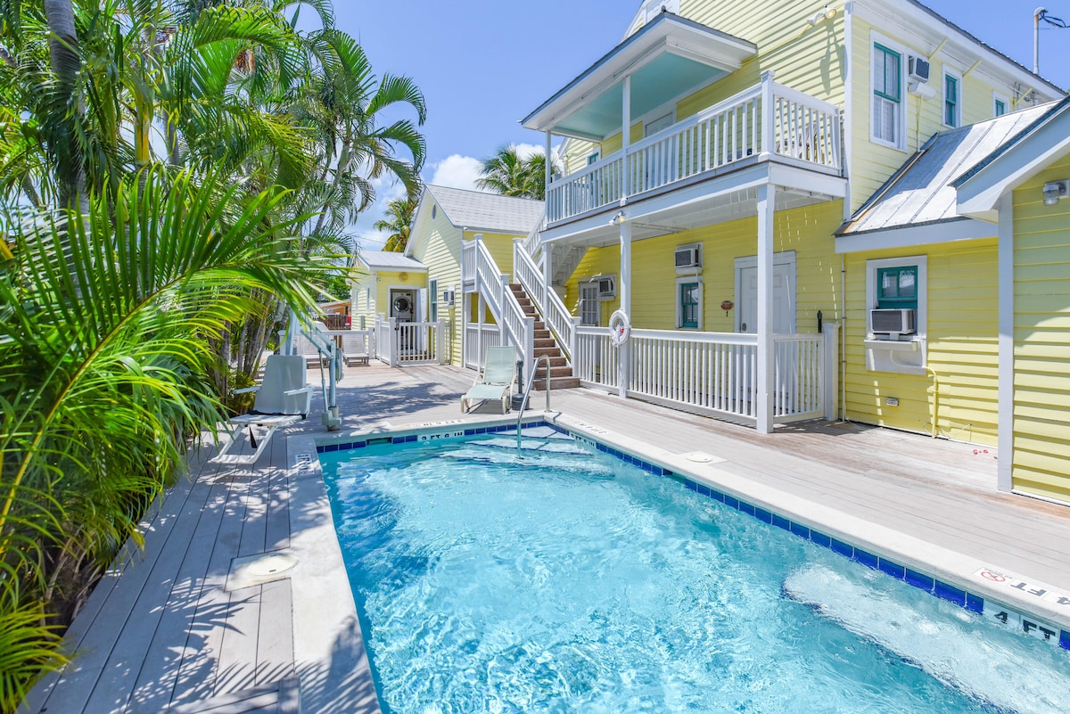A shared heated pool is surrounded by a wooden deck and tropical landscaping, including palm trees. The bright yellow exterior of the two-story building complements the inviting outdoor space. A safety ring is visible on the pool's edge, indicating a family-friendly environment.