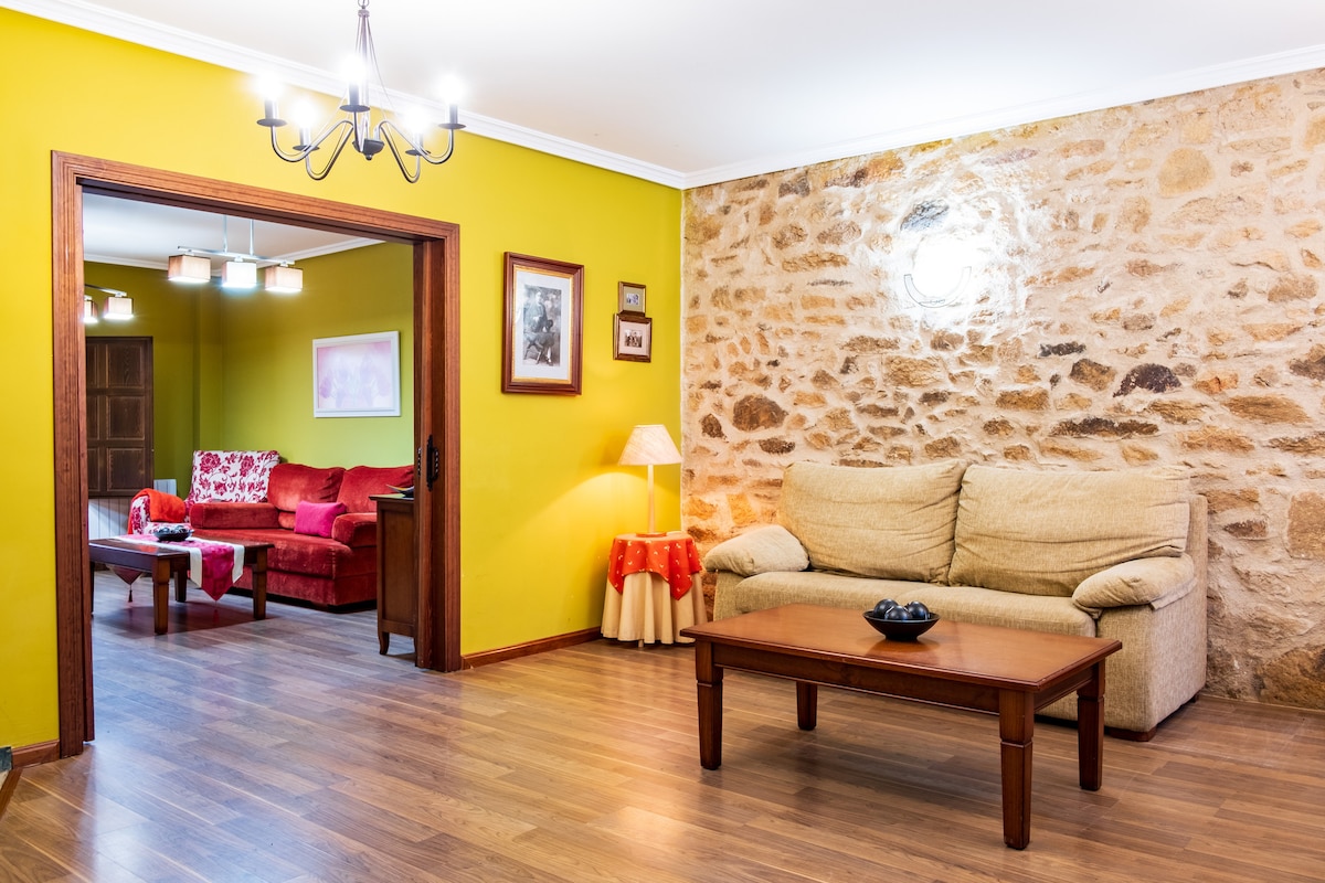 A living room area features a comfortable beige sofa and a wooden coffee table with a decorative bowl. The walls are painted a bright green, contrasting with the exposed stone wall. A doorway leads to a second seating area with a red sofa set, creating a cozy environment.