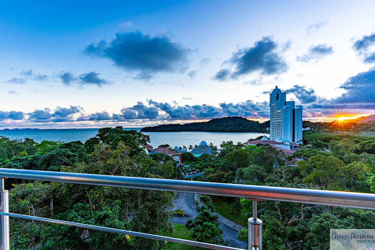 A panoramic view reveals lush green trees and the serene coastline in the distance, with the sun setting near the horizon. Tall buildings are visible along the shoreline, and a clear sky showcases soft clouds. A railing frames the scene, enhancing the outlook.