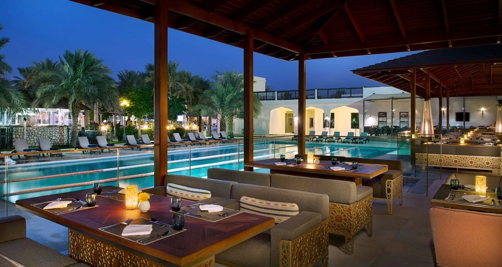 An open-air dining area features multiple wooden tables with comfortable seating, complemented by softly glowing candlelight. Relaxed lounge chairs are situated beside a sparkling pool, bordered by palm trees, creating a serene ambiance in the evening light.
