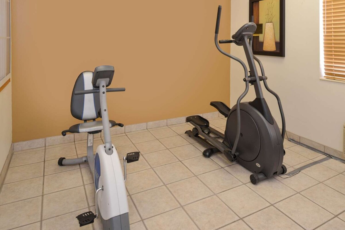 A small exercise area is equipped with a stationary bike and an elliptical machine, situated on tile flooring. The walls are painted a soft, warm hue, and a framed piece of wall art adds a subtle decorative touch.