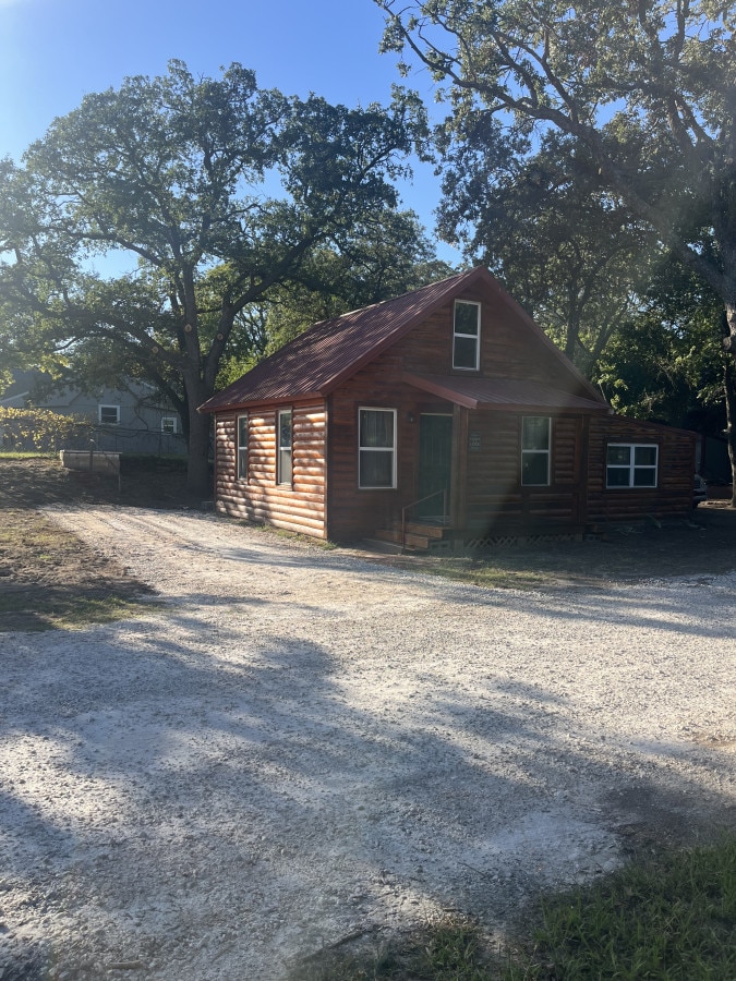 A quaint cabin is showcased surrounded by mature trees, featuring rustic log siding and a welcoming front porch. A gravel pathway leads to the entrance, enhancing the natural setting. The image captures a bright sky, indicating a sunny atmosphere.