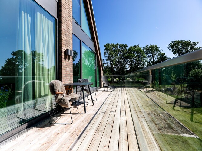 A spacious terrace is featured, showcasing large glass panels and wooden flooring. Two chairs with soft covers and a small table are positioned, providing a place to relax. The tranquil garden view is complemented by lush greenery in the background.