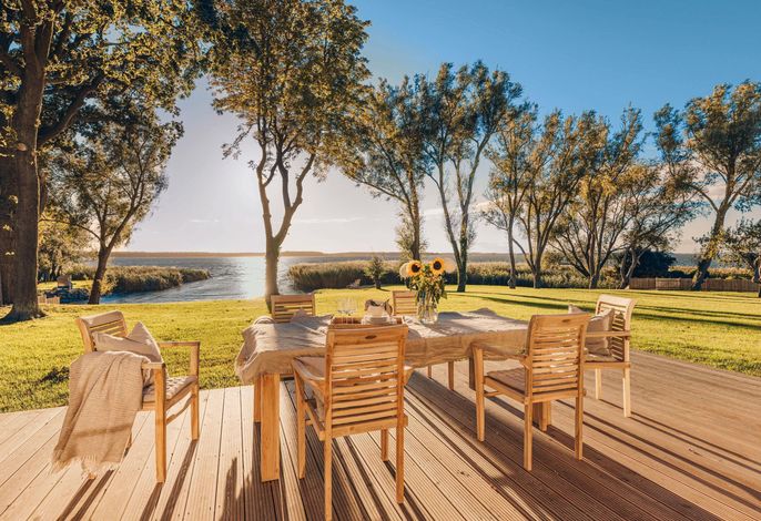 A spacious wooden terrace is shown, furnished with a dining table surrounded by six wooden chairs. Bright sunshine filters through the surrounding trees, providing a view of the water beyond. A vase with sunflowers adds a touch of nature to the setting.