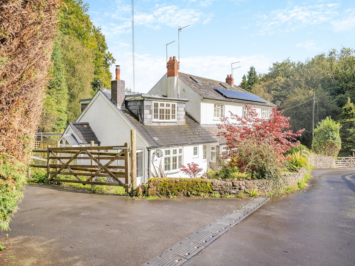 A charming countryside cottage is nestled amidst greenery, featuring white walls and a slate roof. Flowering shrubs and a wooden gate add to its rustic appeal. The surrounding landscape is enhanced by tall trees and a clear blue sky, providing a peaceful rural atmosphere.