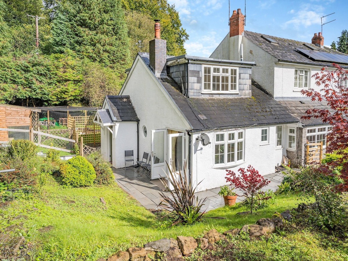 The exterior of the cottage showcases a charming white façade with slate roofing. A patio area is visible, featuring potted plants and seating options. Lush greenery surrounds the property, complemented by a wooden fence and an inviting garden space.