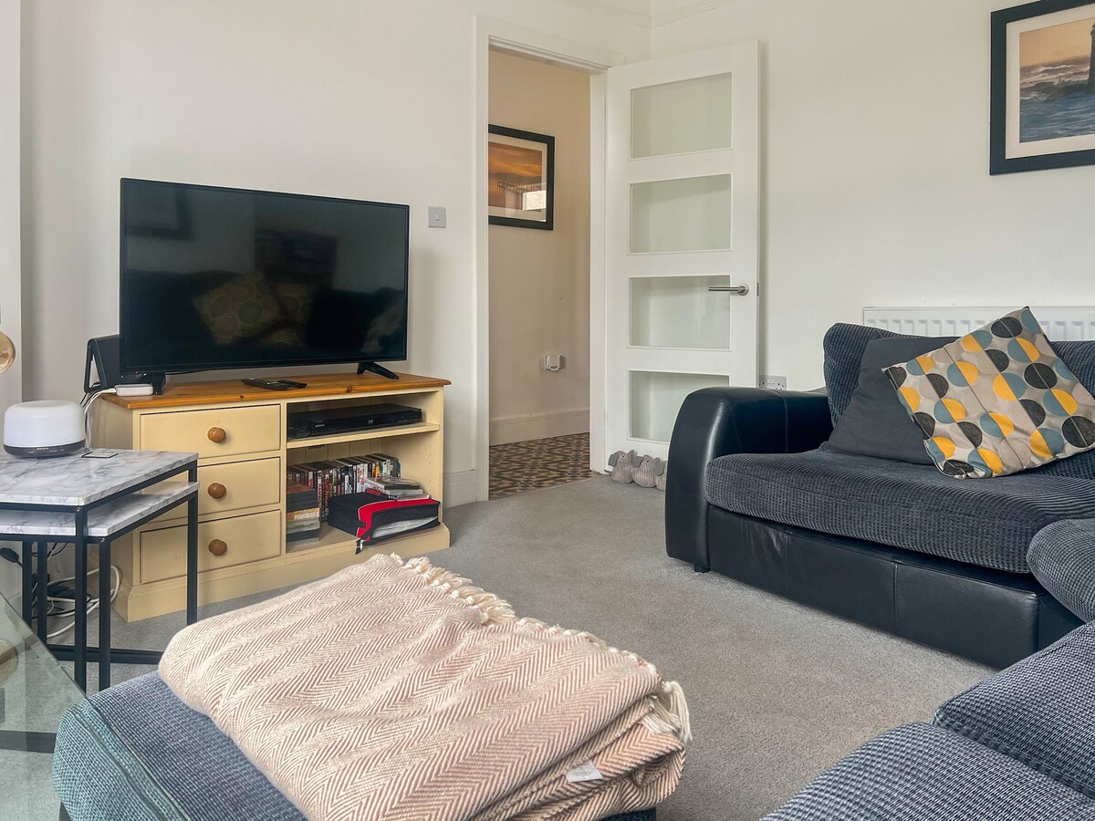 A comfortable living room features a large Smart TV on a wooden console, complemented by a cozy black sofa and a grey textured armchair. A soft throw blanket is draped over a glass coffee table, while a doorway leads into a neighboring room.