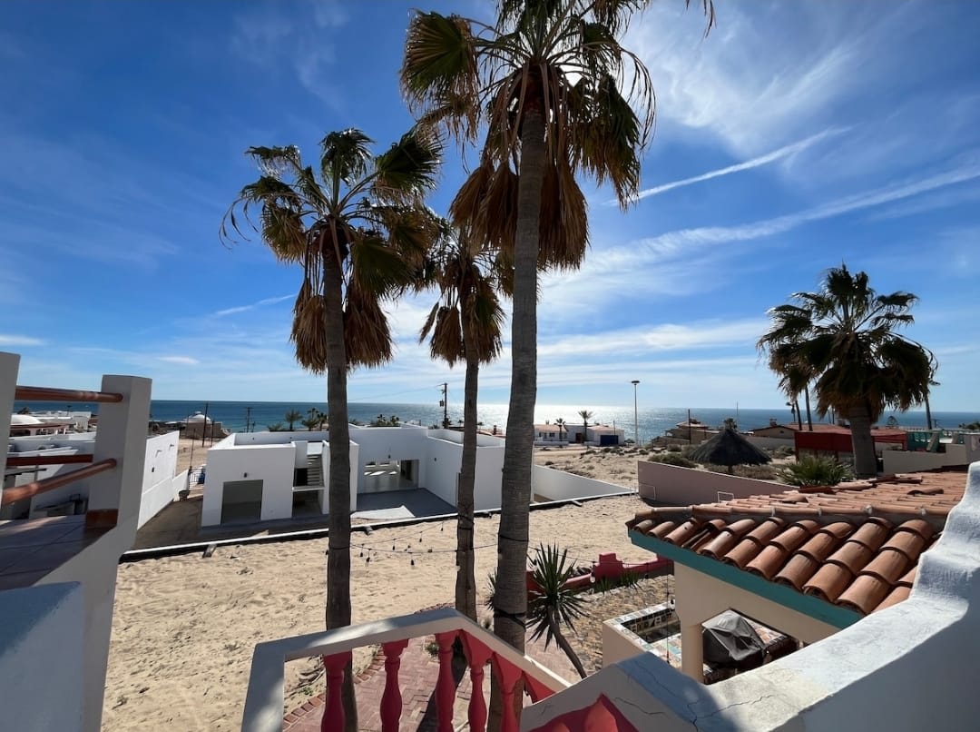 A view from a balcony showcases swaying palm trees overlooking the beach. The ocean's horizon is visible in the distance, with sandy areas and beachgoers spotted along the shore. Nearby structures feature white walls and terracotta rooftops, enhancing the coastal atmosphere.